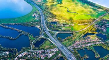 A road and roundabout seen from above, cutting through sunlit green fields and flooded plains