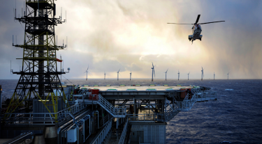 Image shows a helicopter flying above an offshore station with offshore wind turbines in the background